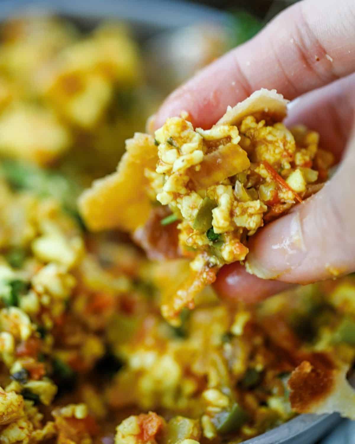 A hand holding a piece of roti topped with Paneer Bhurji and vegetables.